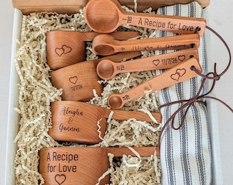 May include: A set of wooden measuring cups and spoons in a cardboard box with a striped cloth. The items are engraved with the text A Recipe for Love, Aleigha & Gannon, 11/7/26, and heart symbols. The spoons include 1 TBS, 1 TSP, 1/2 TSP, and 1/4 TSP.