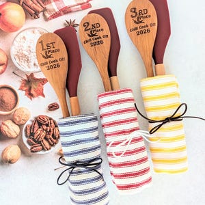 May include: Three wooden spoons and maroon silicone spatulas bundled with striped kitchen towels. The spoons are engraved with 1st, 2nd, and 3rd Place Chili Cook Off 2026. The towels are blue, red, and yellow striped, each tied with a thin cord.