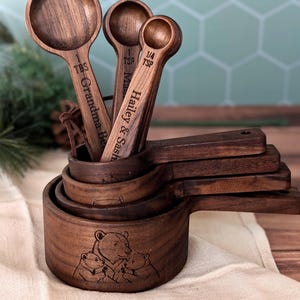 May include: A set of dark brown wooden measuring cups and spoons. The largest cup features an engraved bear family illustration. The handles are engraved with text including 1 TBS, 1 TSP, 1/4 TSP, Grandma Be, and Hailey & Sash.