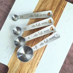 May include: A set of four silver-toned metal measuring spoons with round bowls and flat handles. Each handle is engraved with a leaf sprig, the text Est. 2023, and a heart-shaped cutout. The spoons are labeled 1/4 TSP, 1/2 TSP, 1 TSP, and 1 TBS.