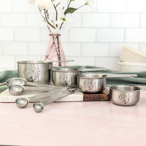May include: A set of stainless steel measuring cups and spoons featuring etched floral heart designs. The cups and spoons have long handles with visible measurement markings, showcasing a metallic silver finish and detailed botanical engravings.