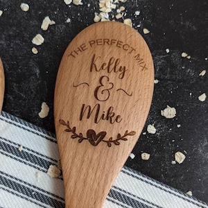 May include: A light brown wooden spoon featuring the laser-engraved text THE PERFECT MIX, Kelly & Mike, and a small heart icon surrounded by leafy branches. The spoon has a smooth finish and a rounded bowl shape with visible wood grain.