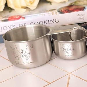 May include: A set of four stainless steel measuring cups of varying sizes, each featuring custom laser-engraved designs and text. The engravings include illustrations of hands, hearts, and the phrases Scott & Alana, Est. 9.14.2021, Wishing you love, and beyond measure.