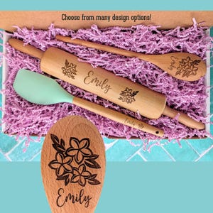 May include: A wooden rolling pin, a wooden spoon, and a light green silicone spatula nestled in purple paper shreds. The rolling pin and spoon are engraved with the name Emily and a floral design. The text Choose from many design options is visible.