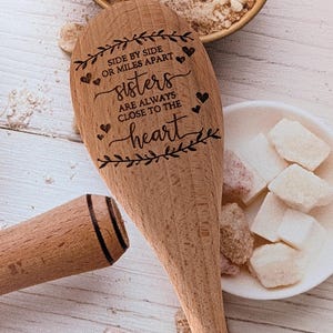 May include: A light brown wooden spoon engraved with the text SIDE BY SIDE OR MILES APART sisters ARE ALWAYS CLOSE TO THE heart, surrounded by decorative leaf motifs and small hearts, positioned near a bowl of sugar and white cubes.