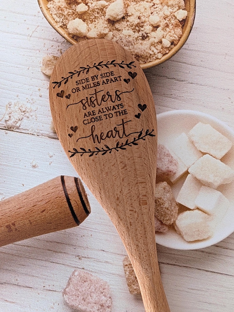 May include: A light brown wooden spoon engraved with the text SIDE BY SIDE OR MILES APART sisters ARE ALWAYS CLOSE TO THE heart, surrounded by decorative leaf motifs and small hearts, positioned near a bowl of sugar and white cubes.