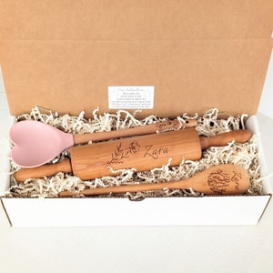 May include: A white box with a pink silicone heart-shaped spatula, a wooden rolling pin with the name "Zara" engraved on it, and a wooden spoon with the name "Zara" engraved on it. The box is filled with white shredded paper.