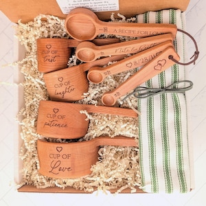 May include: A set of wooden measuring cups and spoons in a box with a green and white striped towel. The cups are engraved with phrases like Cup of Love, Cup of Patience, Cup of Laughter, and Cup of Trust. The spoons feature text and dates.