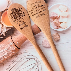 May include: Two light-colored wooden spoons with laser-engraved text and designs. One spoon features a house illustration and the text The Smith Family. The other spoon reads NEW HOME new adventures NEW memories Est. 2026.