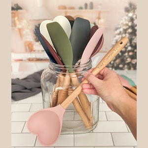Puede incluir: Colecci&oacute;n de esp&aacute;tulas de silicona con forma de coraz&oacute;n en varios colores, incluyendo rosa, verde y blanco, con mangos de madera. Una esp&aacute;tula se sostiene, mostrando el texto "Your Custom Text" en el mango. Las esp&aacute;tulas se guardan en un tarro de cristal transparente.