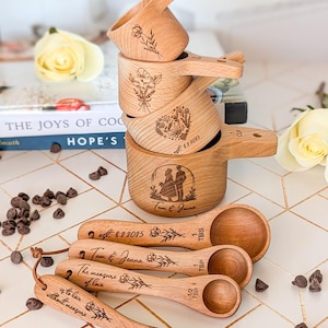 May include: A stack of wooden measuring cups and spoons with engraved floral designs and text. The cups and spoons are light brown. The text includes names and dates. Chocolate chips are scattered around the items.