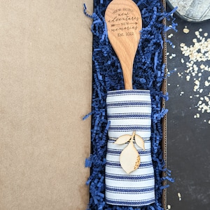 May include: A wooden spoon with the text "New Home, new adventures, new memories, Est. 2022" engraved on it, wrapped in a blue and white striped tea towel and placed in a blue shredded paper-filled gift box.