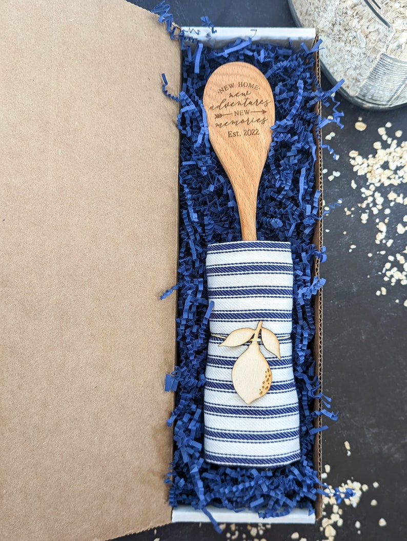 May include: A wooden spoon with the text "New Home, new adventures, new memories, Est. 2022" engraved on it, wrapped in a blue and white striped tea towel and placed in a blue shredded paper-filled gift box.