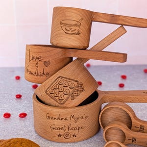 May include: A set of light brown wooden measuring cups and spoons. The cups feature laser-engraved illustrations of a mixing bowl, heart-shaped cookies, and the text Love & Grandkids and Grandma Myra's Secret Recipe. The spoons are labeled 1 TBS, 1 TSP, 1/2 TSP, and 1/4 TSP.