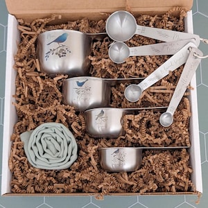May include: A set of four stainless steel measuring cups and four measuring spoons with blue bird illustrations and text including "Mom," "you are a treasure," "& loved beyond," and "all measure" nestled in a box with brown paper filler.