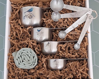 May include: A set of four stainless steel measuring cups and four measuring spoons with blue bird illustrations and text including "Mom," "you are a treasure," "& loved beyond," and "all measure" nestled in a box with brown paper filler.