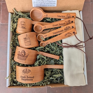 May include: A set of wooden measuring cups and spoons in a cardboard box. The items feature laser-engraved illustrations of flour, jars, and bread, with text including Maria's, Fresh Baked, and Happiness. A beige cloth is tied with a brown cord.