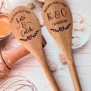 May include: Two light brown wooden spoons with laser-engraved text and designs. One spoon reads THE PERFECT MIX Kate & Caleb with a heart and leaves. The other reads RECIPE FOR LOVE K & C 12/4/2026 with a heart and leaves.