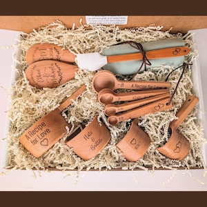 May include: A set of light brown wooden kitchen utensils including spoons, measuring cups, and measuring spoons, all featuring laser-engraved text and heart designs, nestled in a cardboard box filled with shredded paper filler.