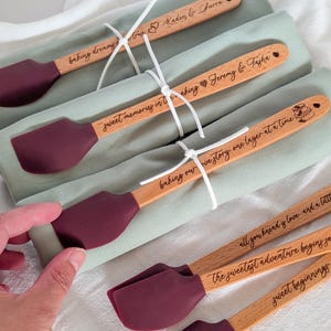 May include: A set of wooden spatulas with dark maroon silicone heads, each featuring laser-engraved text and graphics. Three spatulas are tied with white string atop folded sage green fabric, while two additional spatulas lie below.