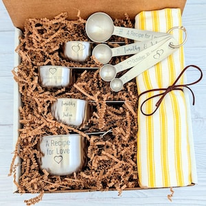 May include: A gift box containing stainless steel measuring cups and spoons with engraved text including A Recipe for Love, Timothy & Julia, and October 10, 2026, alongside a yellow and white striped kitchen towel tied with a brown string.