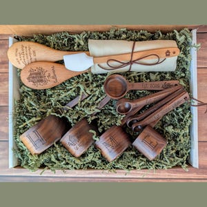 May include: A set of wooden kitchen utensils including two spoons, one with a white silicone tip, and four measuring cups and four measuring spoons. The items feature engraved text including The Sullivans, Est 2025, and letters H, O, M, E.
