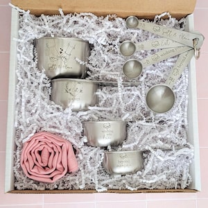 May include: A gift box containing four stainless steel measuring cups and four measuring spoons engraved with text including Scott & Alana, Est 9.14.2025, wishing you love, and beyond measure, alongside a rolled dusty pink fabric napkin on white paper shred.