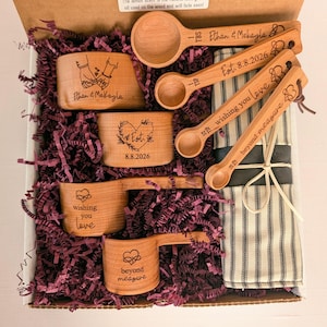 May include: A set of wooden measuring cups and spoons in a box with purple paper filler. The items feature laser-engraved text including Ethan & Mekayla, Est. 8.8.2026, wishing you love, and beyond measure, alongside heart and infinity symbols.