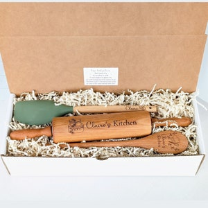 May include: A white gift box filled with brown paper shreds and a set of kitchen tools. The tools include a green silicone spatula, a wooden rolling pin with the text "Claire's Kitchen" and a cake design, a wooden spoon with the text "Claire's Kitchen", and a wooden stirring spoon with the text "Claire's Kitchen".