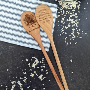 May include: Two wooden spoons with a house design and the text "New Home, new adventures, new memories, Est. 2022, The Workmans" engraved on them. The spoons are on a striped kitchen towel and a dark surface with scattered oats.
