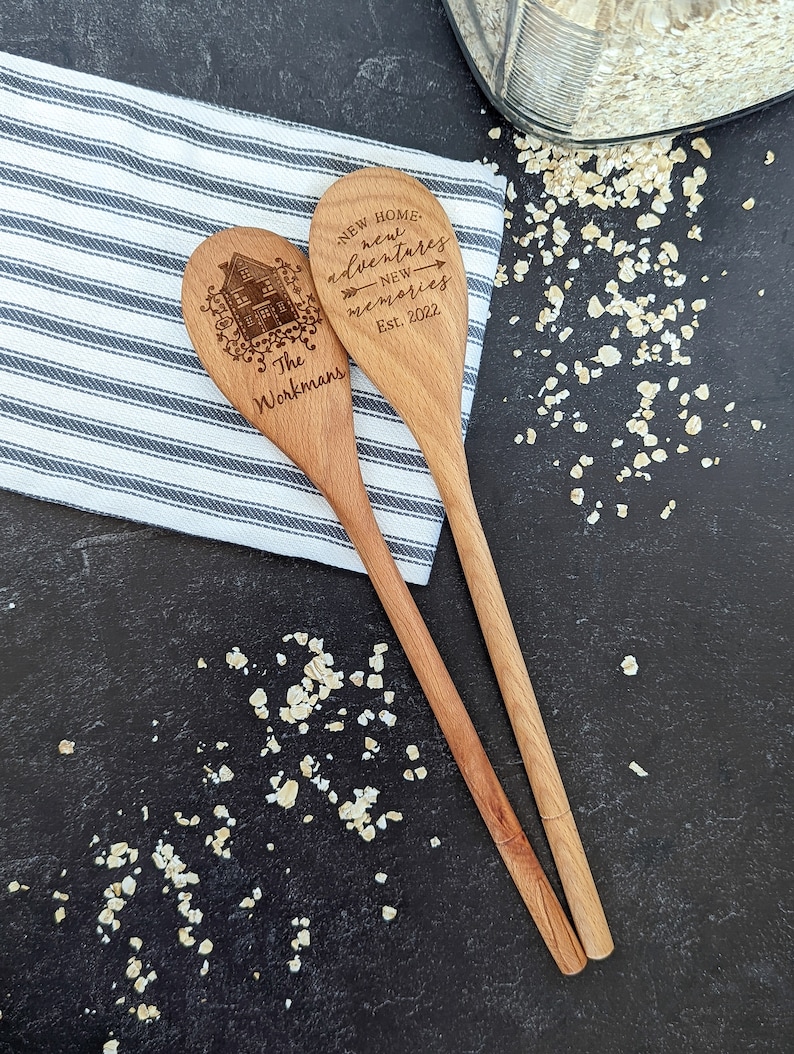 May include: Two wooden spoons with a house design and the text "New Home, new adventures, new memories, Est. 2022, The Workmans" engraved on them. The spoons are on a striped kitchen towel and a dark surface with scattered oats.