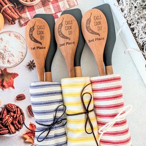 May include: Three wooden spoons with black silicone heads, each engraved with a chili pepper, the year 2026, and the text Chili Cook Off followed by 1st Place, 2nd Place, or 3rd Place. Each spoon is wrapped in a striped kitchen towel tied with string.