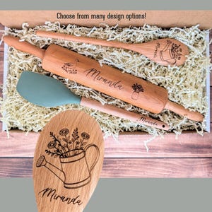 May include: A wooden rolling pin, a wooden spoon, and a sage green silicone spatula nestled in a box with shredded paper. The rolling pin is engraved with the name Miranda and a bird. The spoon features a watering can and flower design.