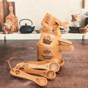 May include: A set of wooden measuring cups and spoons. The cups are engraved with images of baked goods and the words "Fresh Baked Happiness." The spoons are labeled with measurements. A leather cord is attached to the spoons.