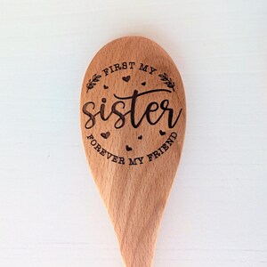May include: A light brown wooden spoon featuring a laser-engraved design. The text reads FIRST MY SISTER FOREVER MY FRIEND in a mix of script and block lettering, surrounded by small heart icons and two leafy branch illustrations.