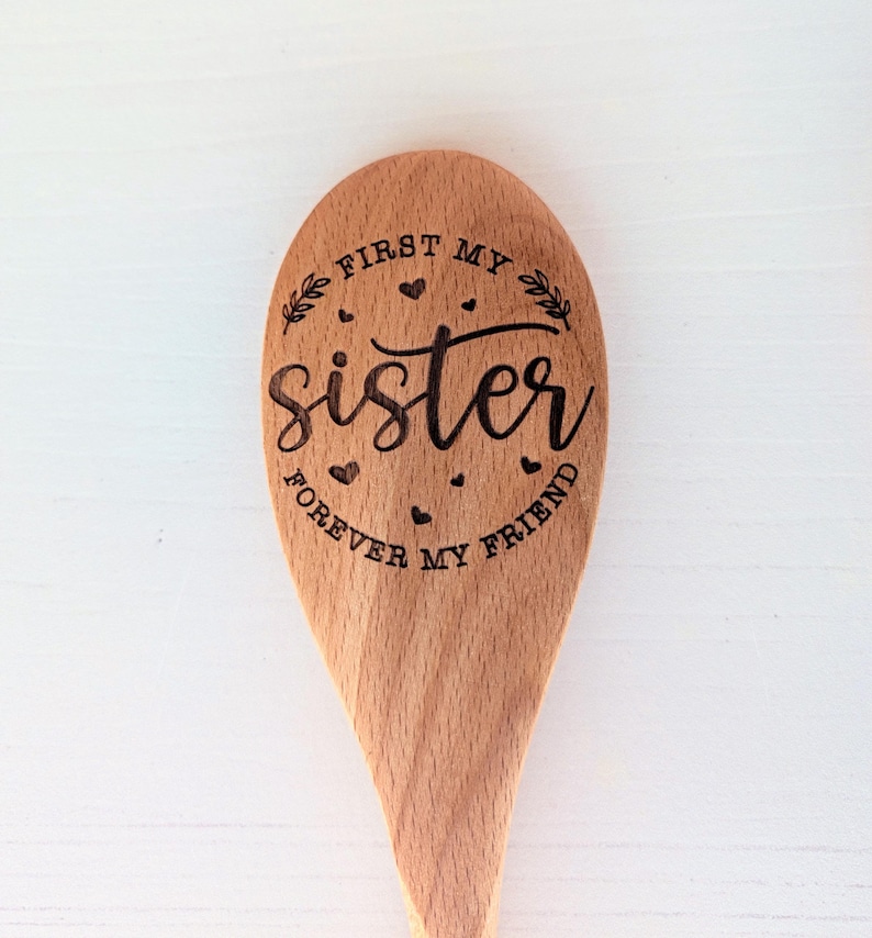 May include: A light brown wooden spoon featuring a laser-engraved design. The text reads FIRST MY SISTER FOREVER MY FRIEND in a mix of script and block lettering, surrounded by small heart icons and two leafy branch illustrations.