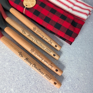 Puede incluir: Cuatro cucharas de cocina de madera con texto grabado "Wishing you Joy" y "Merry Christmas 2025". Las cucharas tienen mangos de madera natural, puntas rojas y cabezas de esp&aacute;tula negras. Un pa&ntilde;o a cuadros rojo y negro y un pa&ntilde;o a rayas est&aacute;n en el fondo.