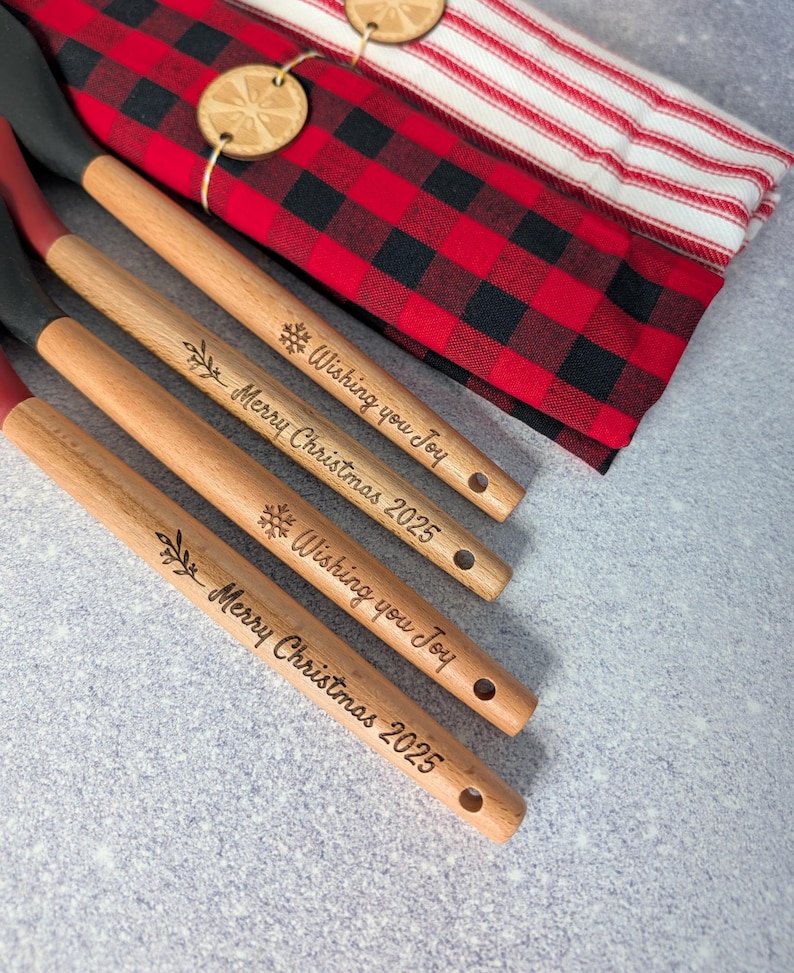 Puede incluir: Cuatro cucharas de cocina de madera con texto grabado "Wishing you Joy" y "Merry Christmas 2025". Las cucharas tienen mangos de madera natural, puntas rojas y cabezas de esp&aacute;tula negras. Un pa&ntilde;o a cuadros rojo y negro y un pa&ntilde;o a rayas est&aacute;n en el fondo.