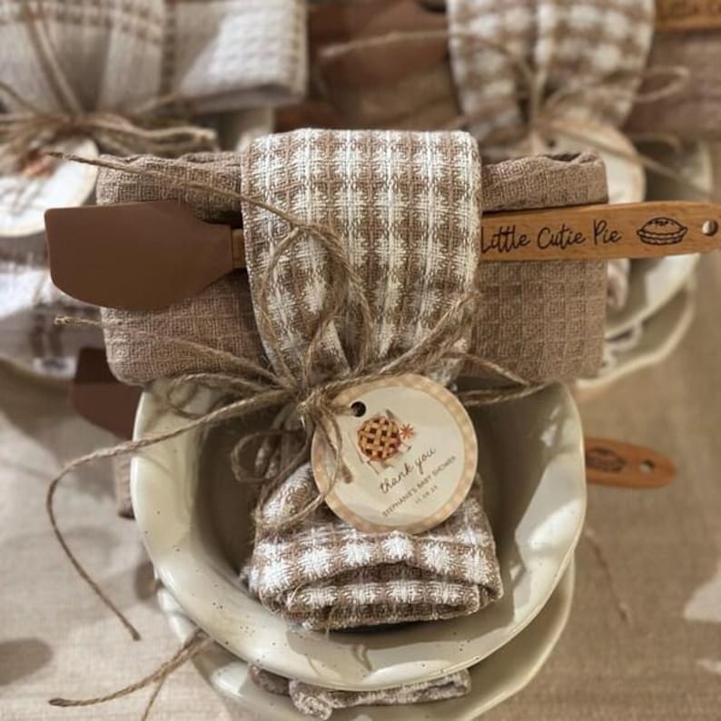 Pu&ograve; includere: Un canovaccio da cucina a nido d'ape a quadri beige e bianchi avvolto attorno a una ciotola in ceramica chiara. Una spatola in legno con la scritta "Little Cutie Pie" e un disegno di una torta &egrave; inserita. Un'etichetta rotonda riporta "thank you".