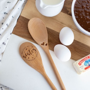 May include: Two light brown wooden spoons with laser-engraved details. One spoon features the text "First my sister, forever my friend" with small hearts, while the other spoon has two small heart engravings. The spoons are placed on a white and wood cutting board.
