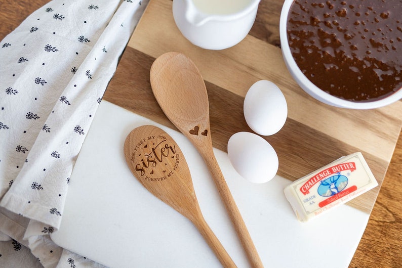 May include: Two light brown wooden spoons with laser-engraved details. One spoon features the text "First my sister, forever my friend" with small hearts, while the other spoon has two small heart engravings. The spoons are placed on a white and wood cutting board.