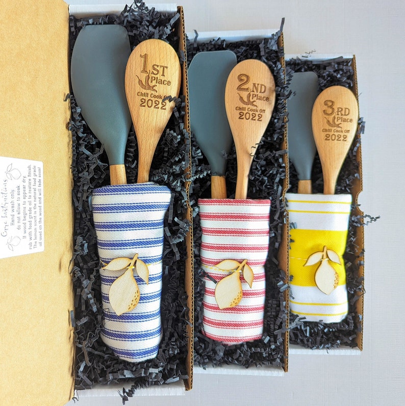 Peut inclure: Trois cuill&egrave;res en bois avec du texte grav&eacute;, "1st Place Chill Cook Off 2022", "2nd Place Chill Cook Off 2022" et "3rd Place Chill Cook Off 2022", sont pr&eacute;sent&eacute;es dans un coffret cadeau avec du papier d&eacute;chiquet&eacute; noir. Chaque cuill&egrave;re est associ&eacute;e &agrave; une spatule en silicone et &agrave; un torchon ray&eacute;.