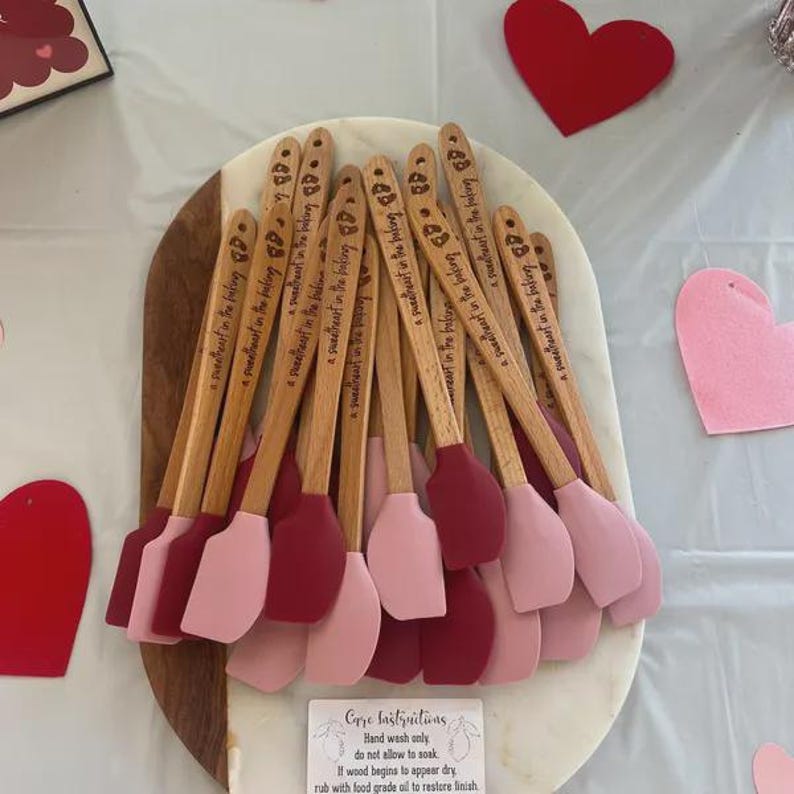 Pu&ograve; includere: Una collezione di spatole con manico in legno e teste in silicone nei toni del rosa e del rosso. Ogni manico &egrave; inciso con il testo 'a sweetheart in the baking'. Un piccolo cartoncino rettangolare in basso fornisce le istruzioni per la cura.