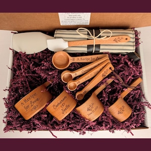 May include: A gift box containing wooden kitchen utensils including measuring spoons and cups engraved with "A Recipe for Love," "Cameron & Joelle," and "June 27, 2026," alongside a white silicone spatula and a striped fabric kitchen towel.
