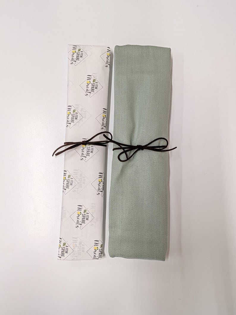 May include: A rectangular sage green fabric item folded and tied with a dark brown cord next to a white paper wrapper featuring the repeating black text A FEW SPARE MOMENTS inside diamond outlines and small yellow floral accents.