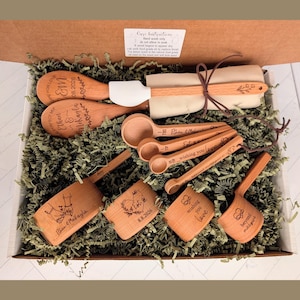 May include: A wooden kitchen utensil set in a cardboard box, including two spoons, four measuring cups, and four measuring spoons. The items feature engraved text and designs, including names, dates, and phrases like 'wishing you love' and 'beyond measure'.