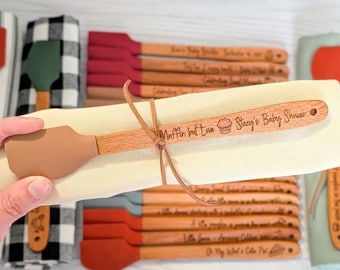May include: Wooden spatulas with brown silicone heads, some with engraved text. One spatula is wrapped in a beige cloth and tied with twine. The text on the spatula reads "Muffin but Love Stacy's Baby Shower". Other spatulas have red and orange silicone heads.