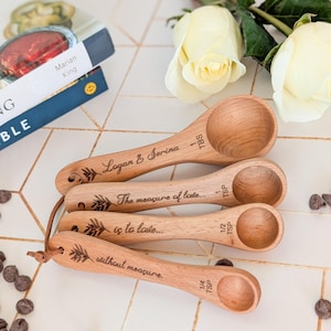 May include: Four wooden measuring spoons with engraved text and floral designs. The spoons are labeled with measurements: 1 TBS, 1/2 TSP, and 1/4 TSP. The text includes "Logan & Serina" and "The measure of love... is to love... without measure."