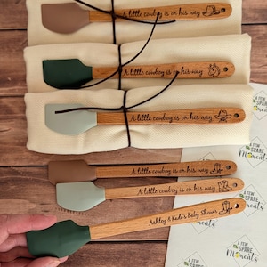 May include: Six wooden-handled silicone spatulas with various colored heads including brown, dark green, and light gray. The handles are engraved with the text A little cowboy is on his way or Ashley & Kade's Baby Shower, accompanied by cowboy-themed icons.