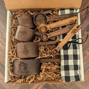 May include: A set of dark brown wooden measuring cups and spoons inside a cardboard box with paper filler. The items feature laser-engraved nature designs and the text The Vigil Cabin. A black and white checkered cloth is included.